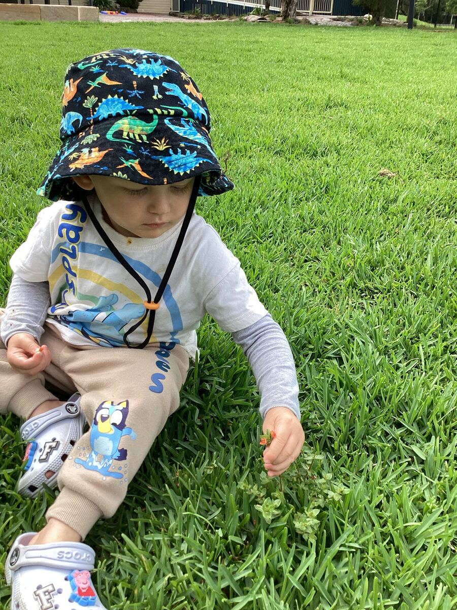 A toddler in a sun hat picks flowers while sitting on grass