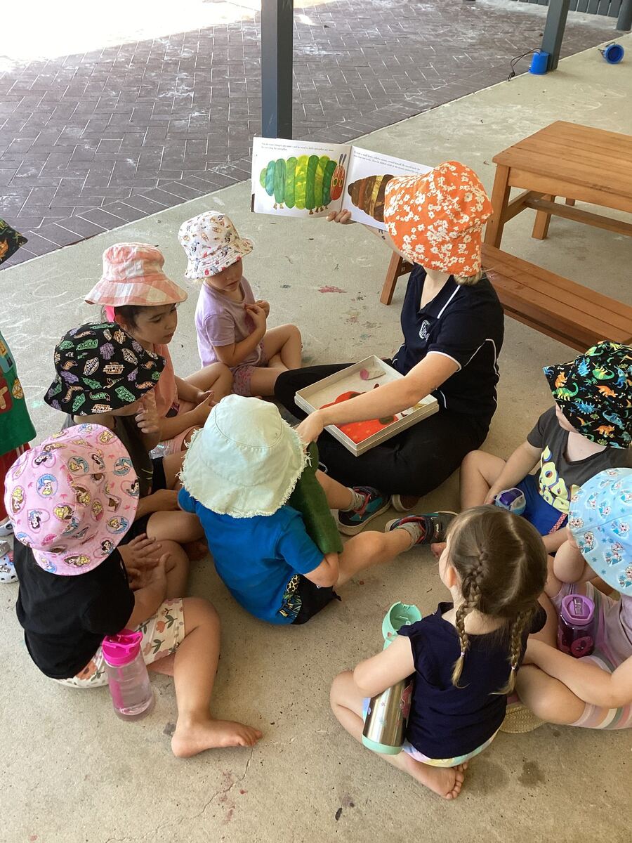 An educator reads to a group of toddlers sitting on the grass outdoors at Explorers