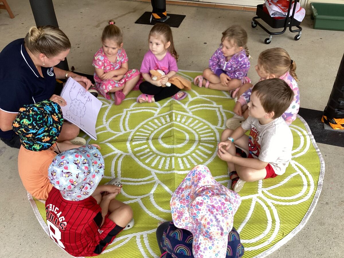 An educator reads to seven children sitting on a circular mat outdoors