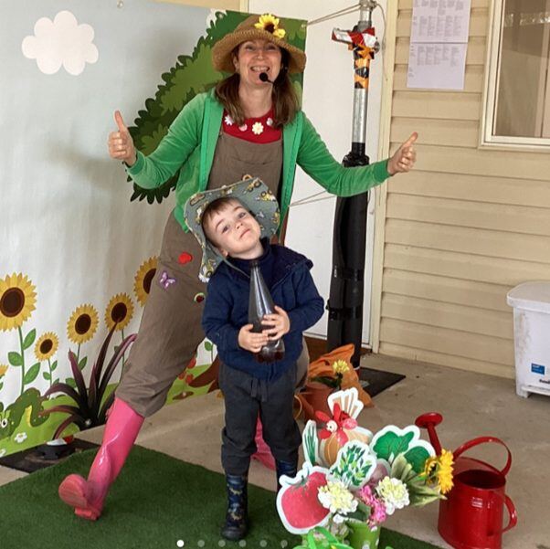 A child and educator both give thumbs up while holding a watering can in the garden